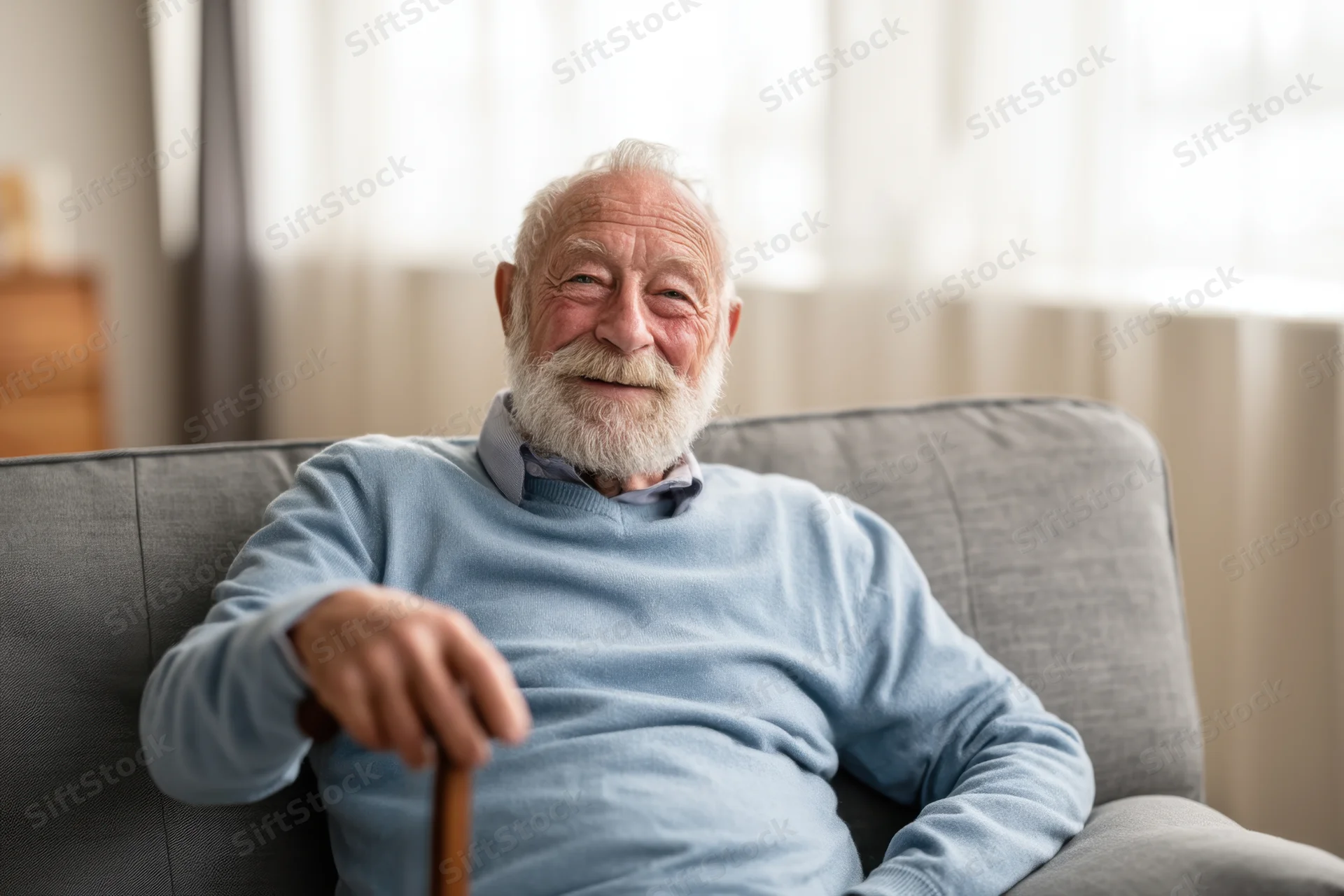 Elderly man with white beard smiling while sitting relaxed on gray couch holding cane in living room