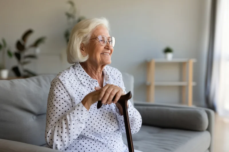Elderly woman smiling while sitting on a modern grey sofa holding a cane in a bright living room
