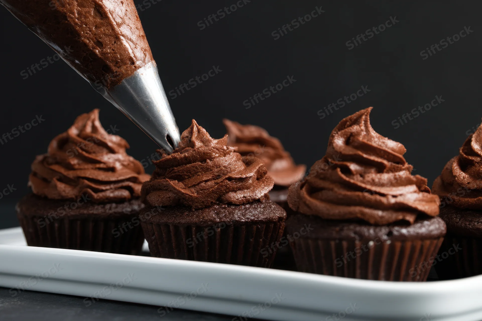 Chocolate cupcakes being decorated with frosting from a piping bag on a white tray