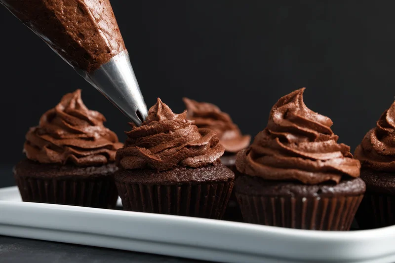 Chocolate cupcakes being decorated with frosting from a piping bag on a white tray