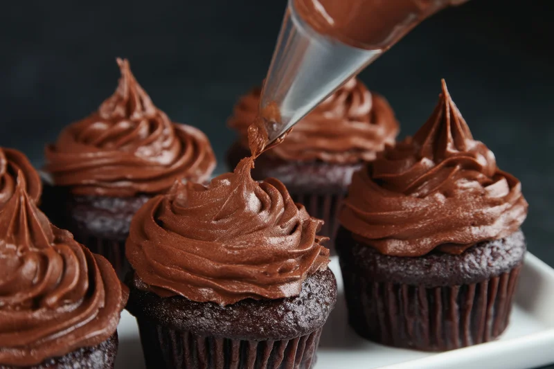 Chocolate cupcakes being decorated with rich chocolate frosting using a piping bag