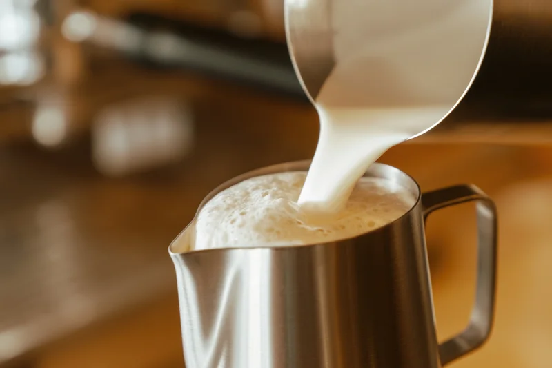 Steamed milk pouring into a stainless steel pitcher on a wooden counter