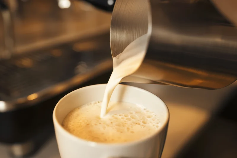 Steamed milk pouring into a white ceramic cup with froth, in a coffee shop setting