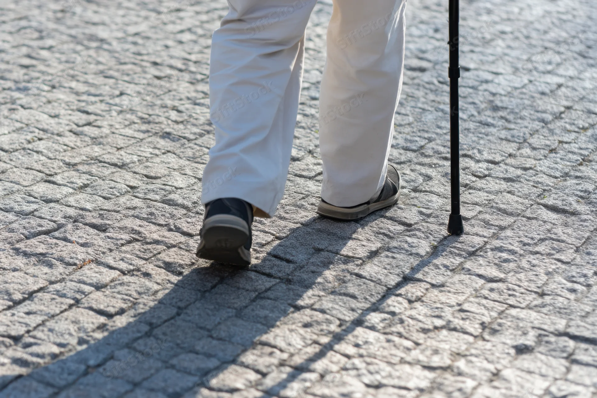 Lower legs of a senior man walking with a cane on a cobblestone pavement