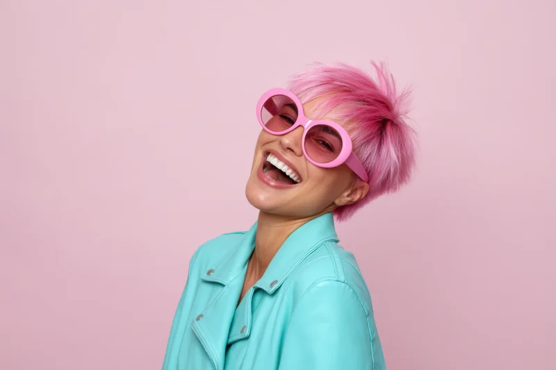 Woman with pink hair and sunglasses laughing in a studio setting wearing a turquoise jacket