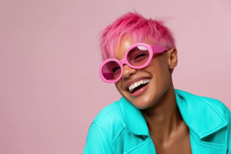 Woman with vibrant pink hair and pink sunglasses smiling in a turquoise jacket against a pink background