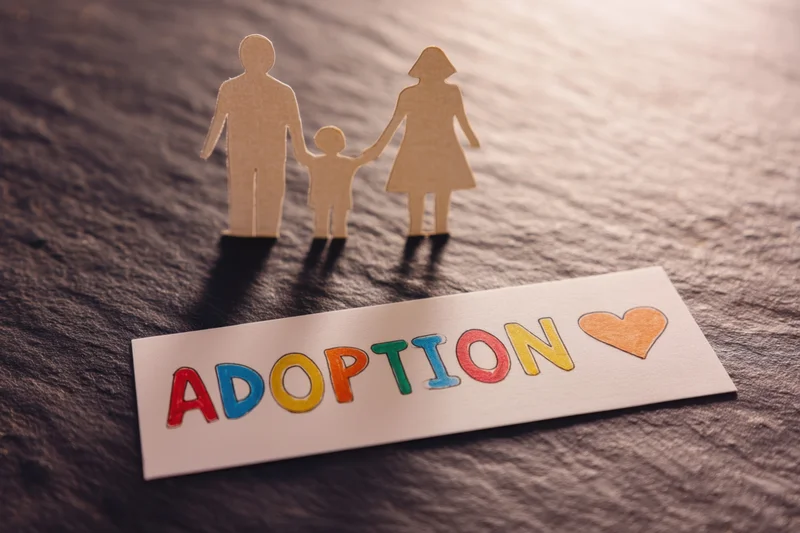 Paper cutout family figures standing beside a colorful adoption sign on a dark textured surface