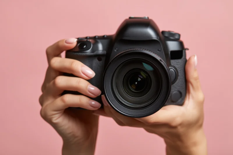 Hands holding a black dslr camera with large lens against pink background