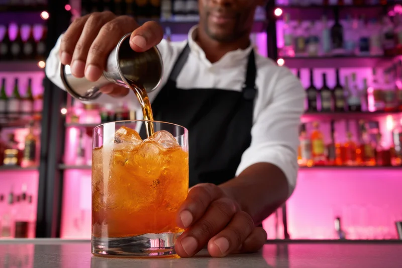 Bartender pouring amber cocktail into glass with ice at bar