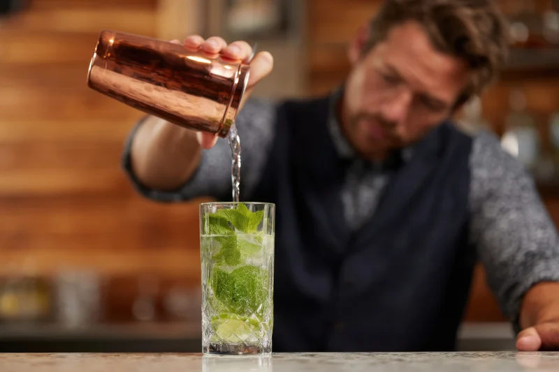 Bartender pouring clear liquid into a glass with mint and lime at a bar
