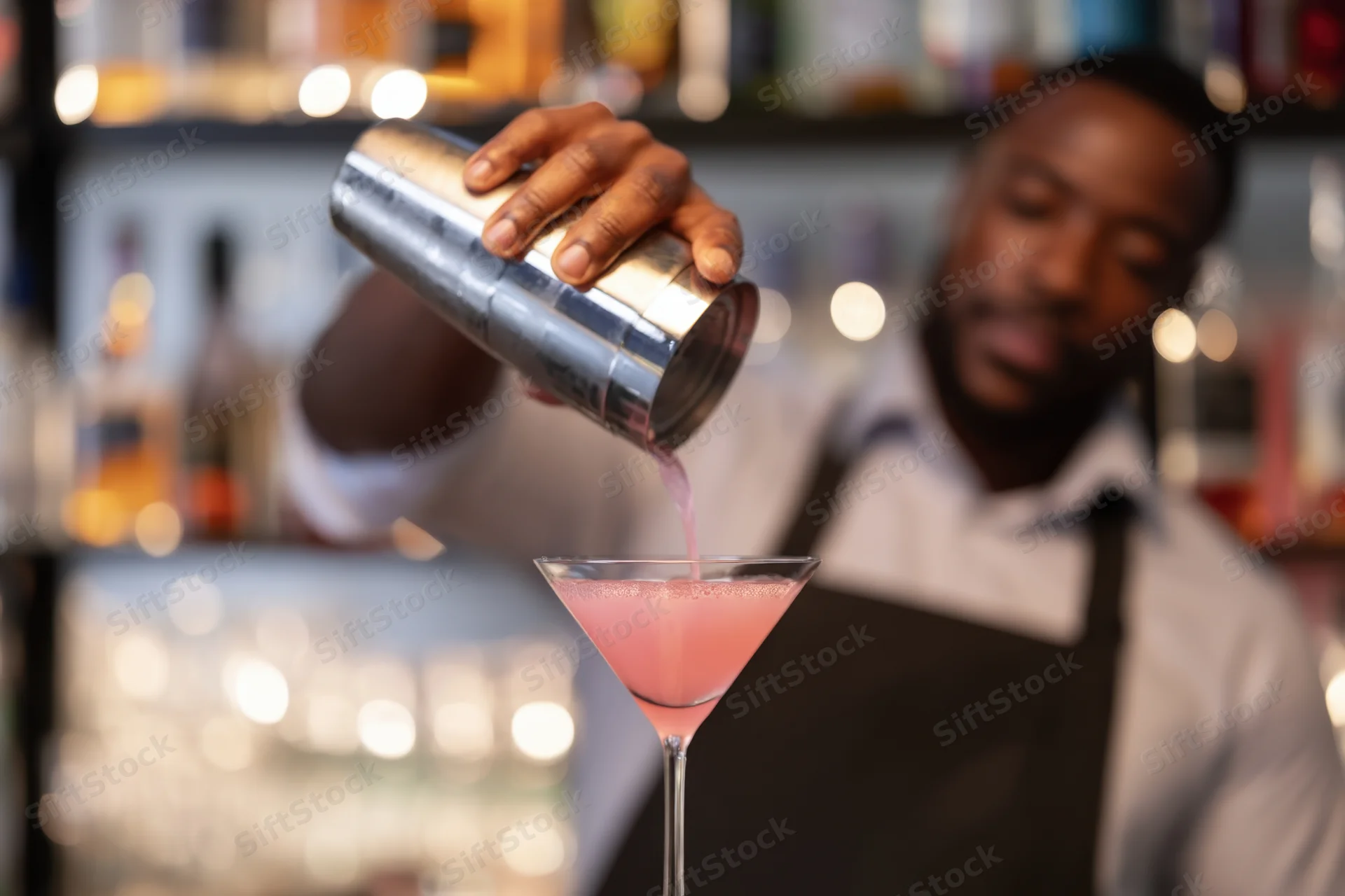 Bartender pouring pink cocktail from stainless steel shaker into martini glass at bar