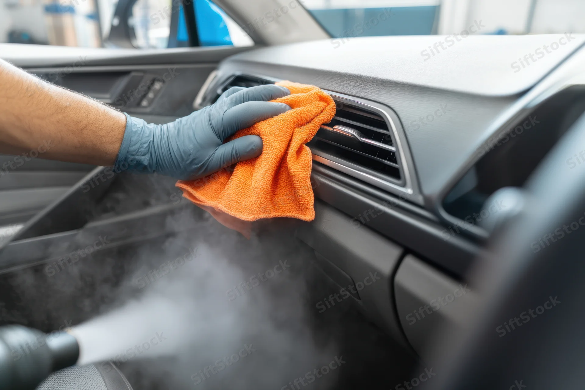 Gloved hand wiping car dashboard vent with orange cloth while steam is applied