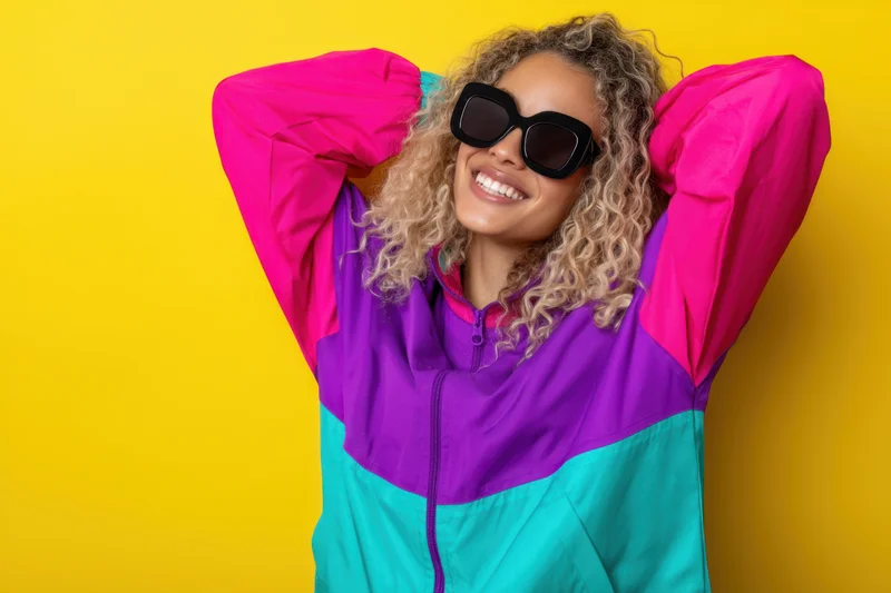 Young woman with curly blonde hair smiling while wearing colorful retro jacket and sunglasses against yellow background