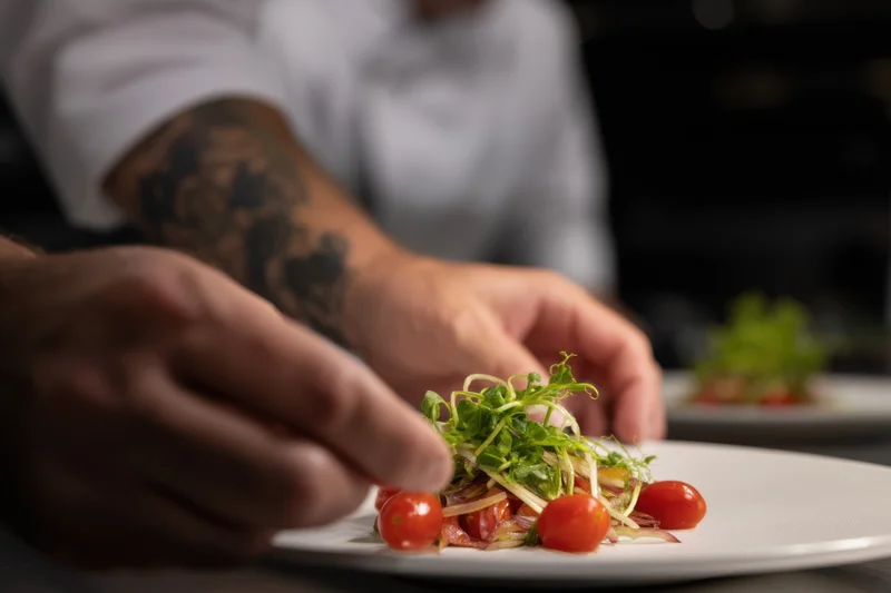 Chef's hands plating a gourmet dish with cherry tomatoes and microgreens on a white plate