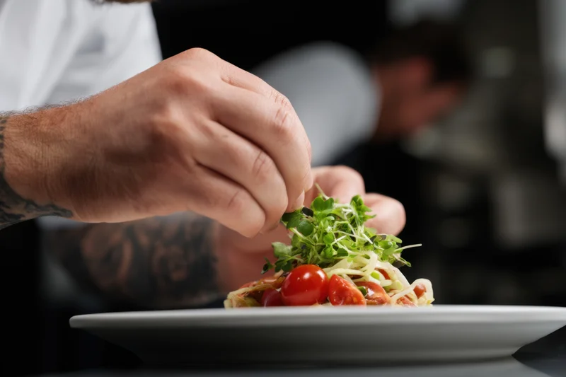 Chef's hands plating a gourmet pasta dish with cherry tomatoes and microgreens on a white plate in a restaurant kitchen