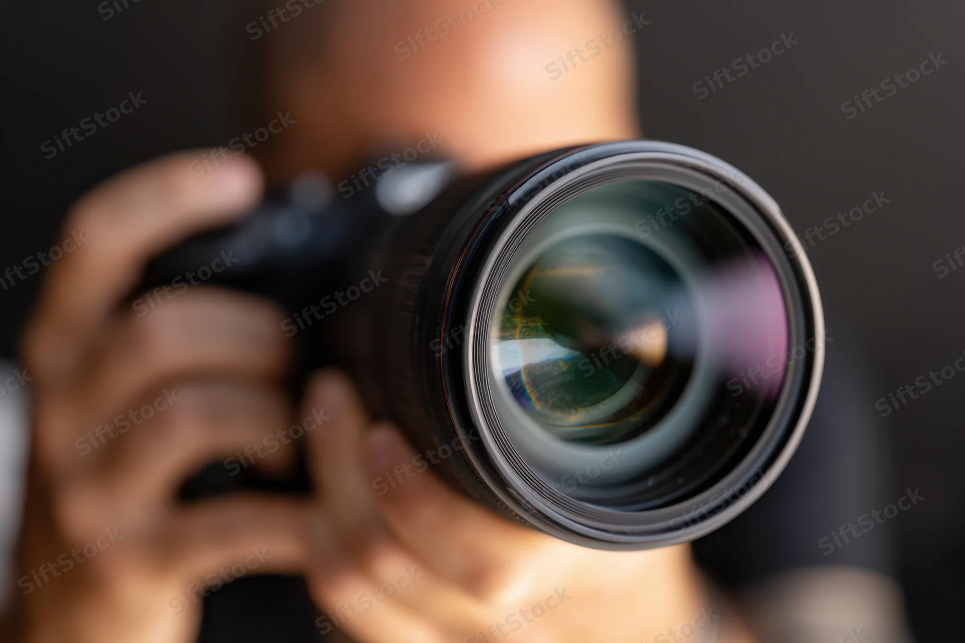 Close-up of a professional DSLR camera held by a photographer with lens in focus, hands gripping body, dark background