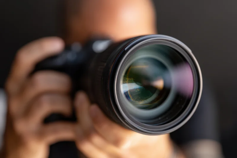 Close-up of a professional DSLR camera held by a photographer with lens in focus, hands gripping body, dark background