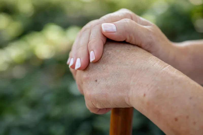 Close-up of an elderly woman's hand being held by a caregiver's hand on a wooden cane outdoors