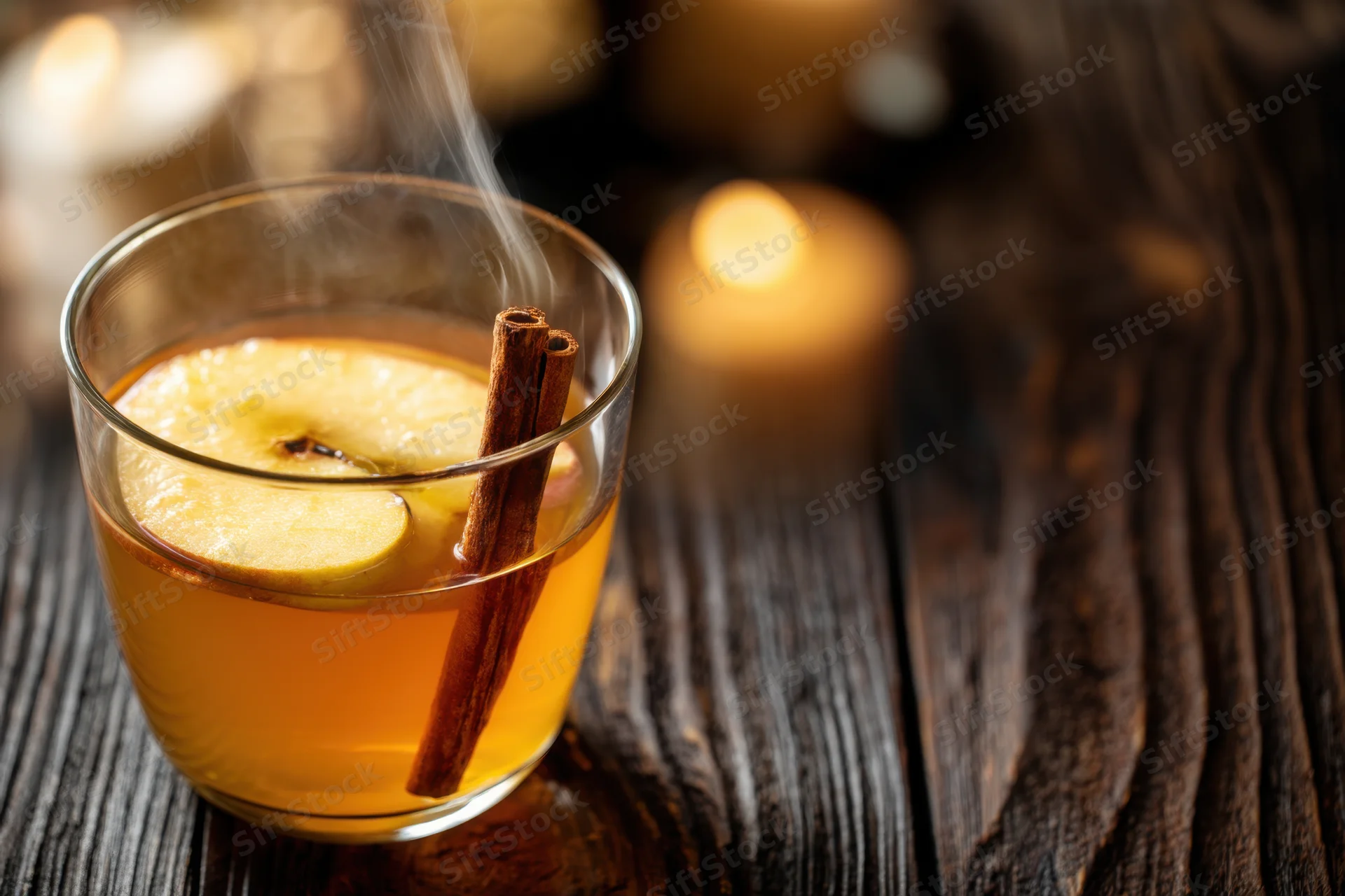 Warm spiced apple cider with apple slice and cinnamon stick in glass on dark wooden table near candlelight