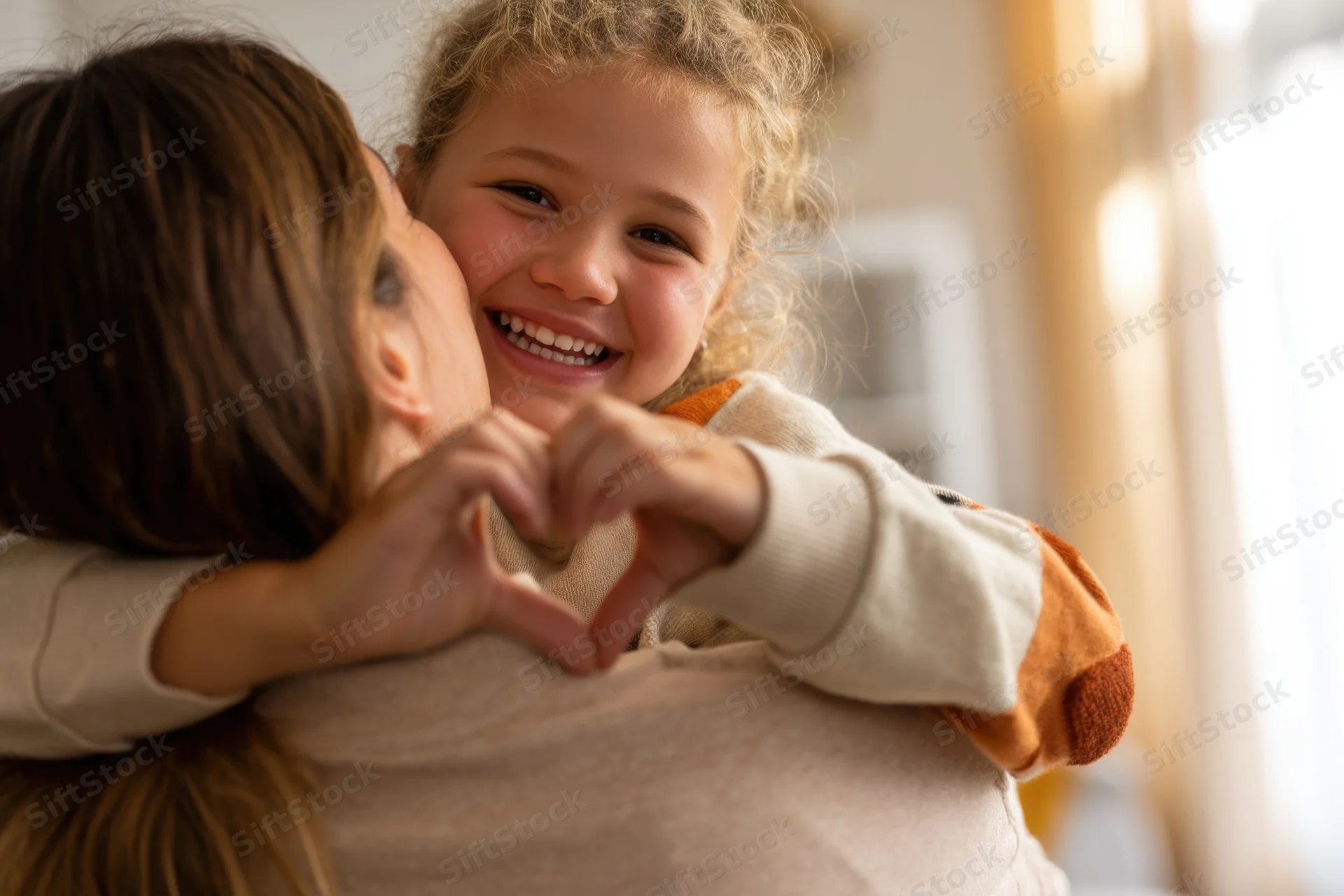Mother hugging daughter who makes heart shape with hands indoors