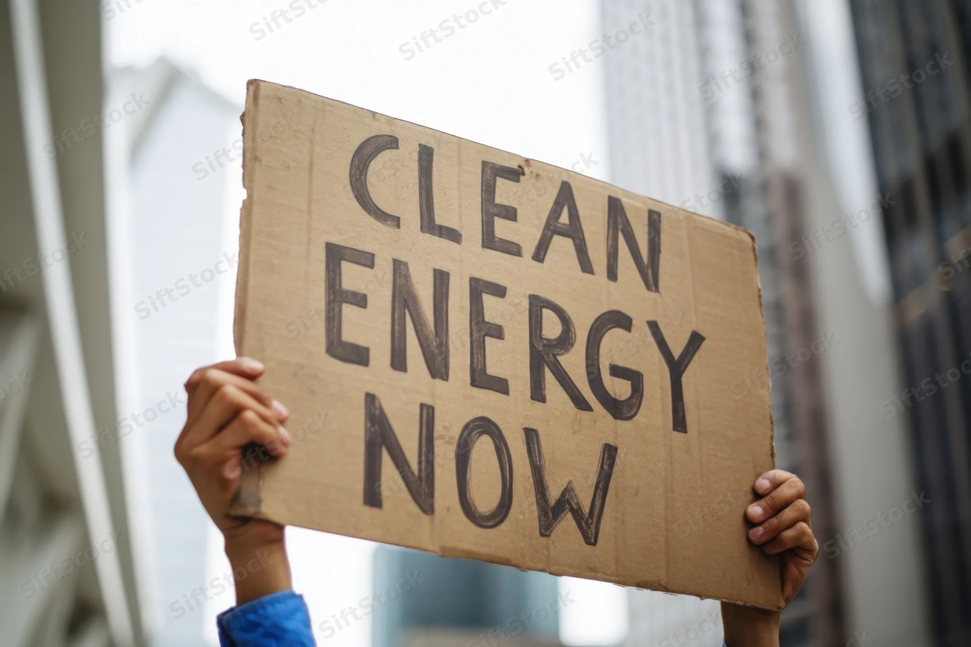 Hands holding a cardboard sign with text clean energy now in an urban setting
