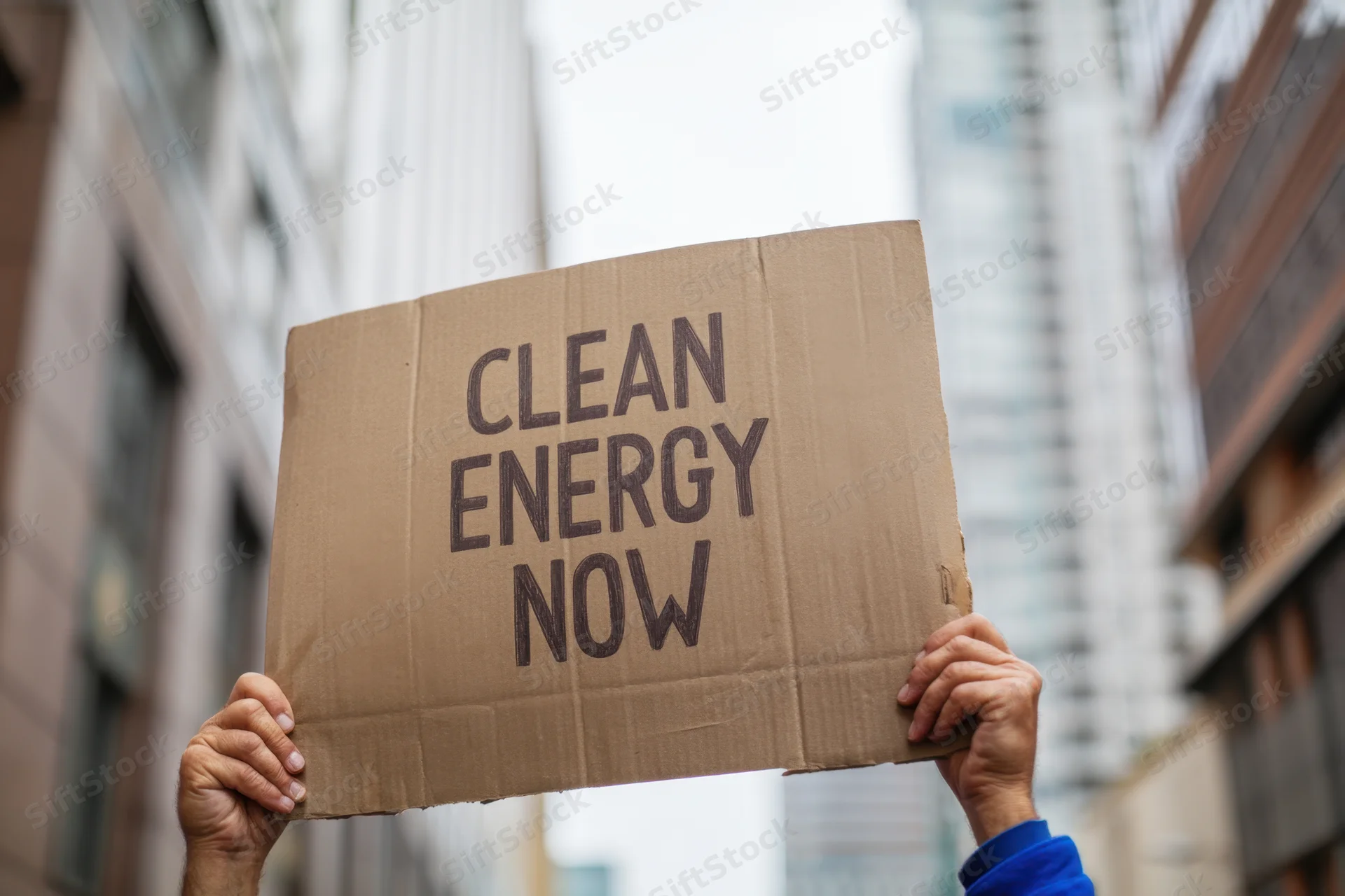 Hands holding a cardboard sign with text clean energy now in an urban street setting