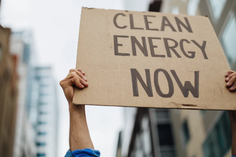 Hands holding a cardboard sign with text clean energy now in an urban setting