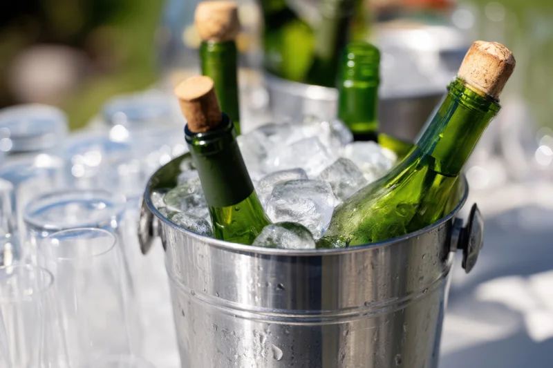 Green wine bottles with corks chilling in a metallic ice bucket on a table outdoors with glasses nearby