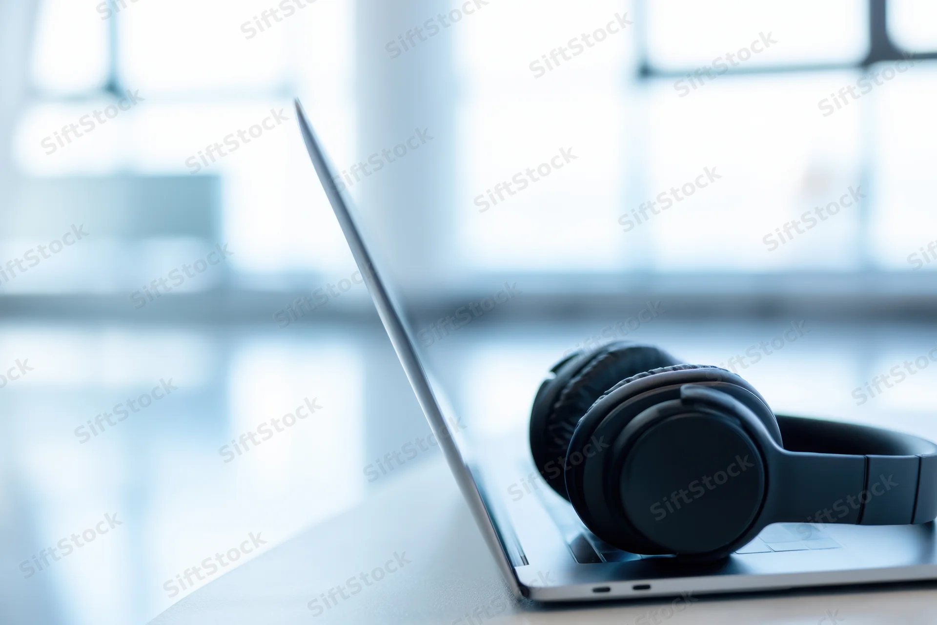 Modern laptop with black over-ear headphones resting on its side on a desk near a window