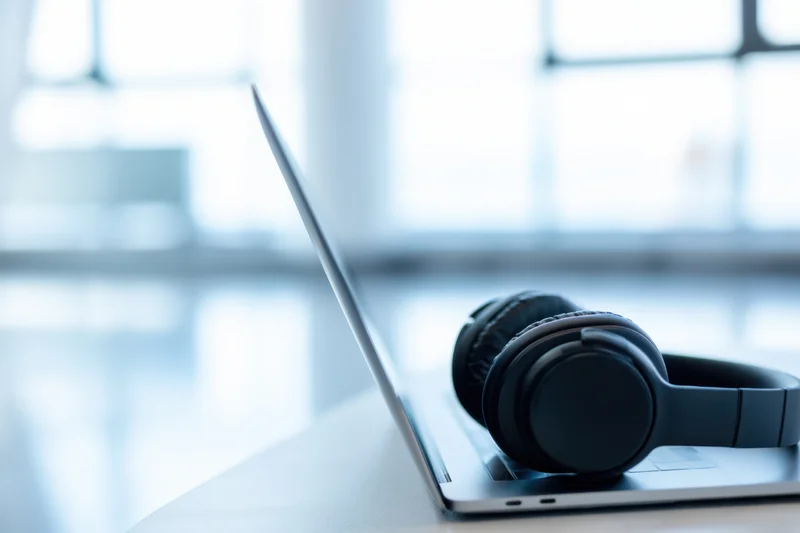 Modern laptop with black over-ear headphones resting on its side on a desk near a window