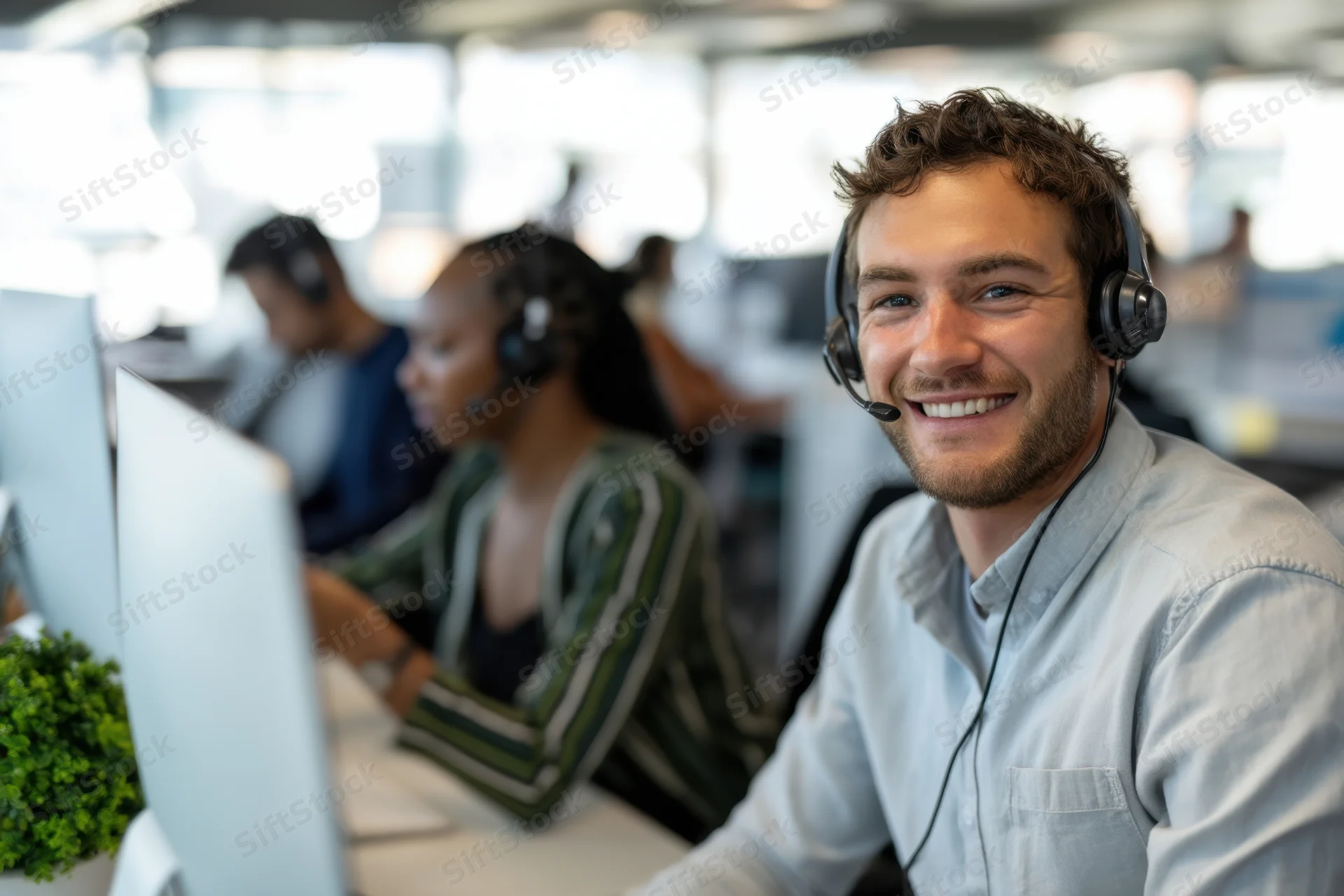 Young man smiling while wearing headset in modern office call center with colleagues at desks