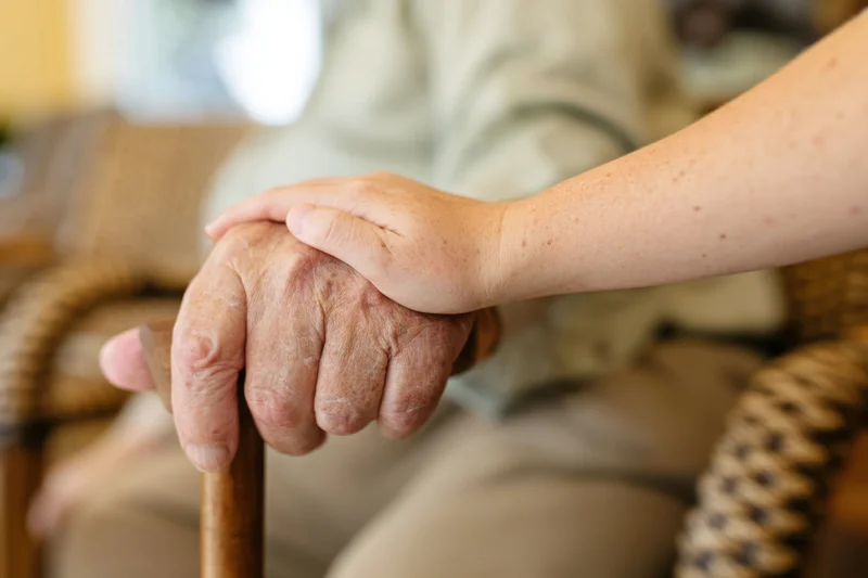 Elderly hand holding a cane with a younger hand resting on top in a seated indoor setting