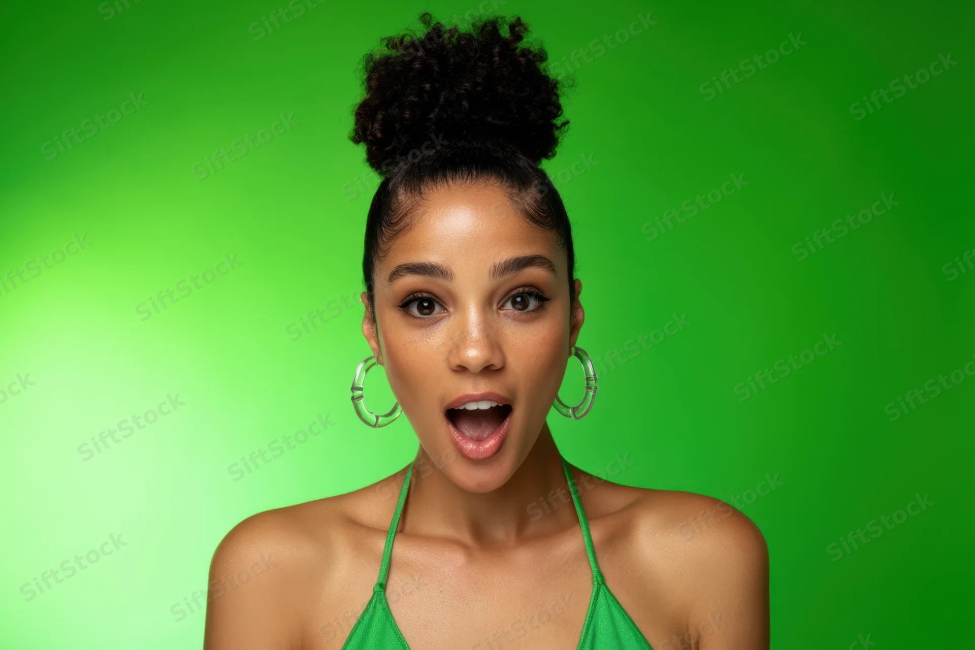 Woman with surprised expression wearing green halter top and hoop earrings against green background