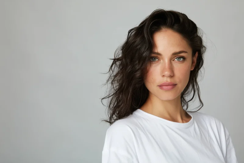Woman with dark wavy hair and intense expression wearing white tshirt in studio setting