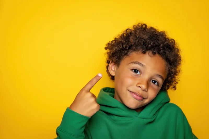 Young boy pointing to his head with a playful smile against a yellow background wearing a green hoodie