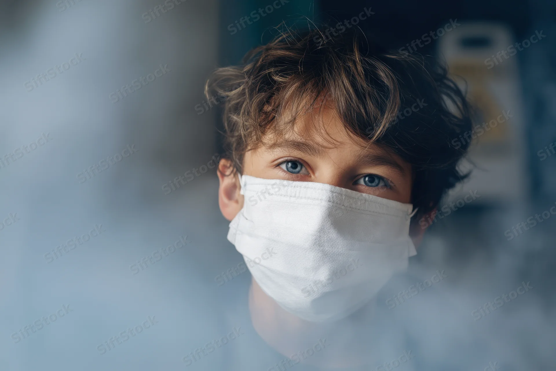 Young boy wearing white face mask with tousled brown hair, looking through window with blue eyes visible above mask