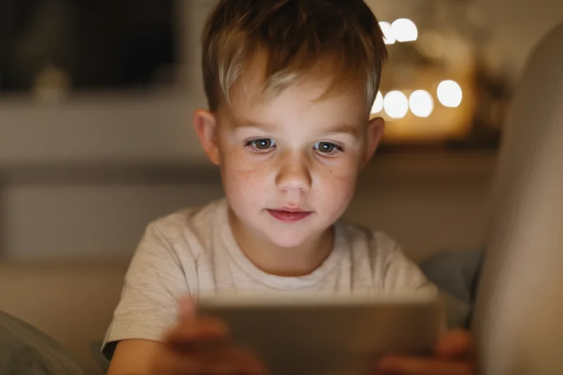 Young boy sitting on a soft couch holding a tablet device in a dimly lit living room