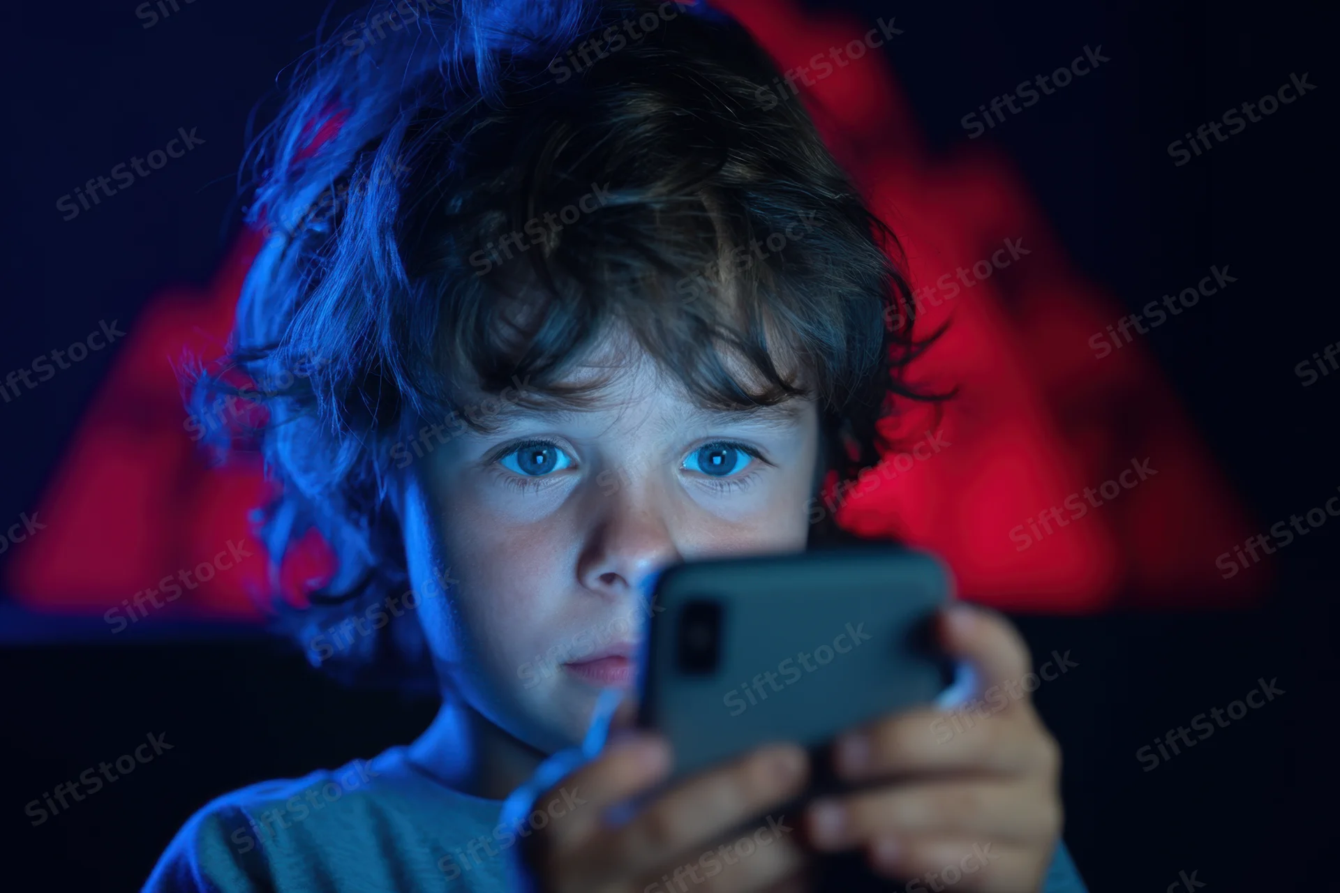 Young child with tousled brown hair and bright blue eyes concentrating on a smartphone screen in a dark room lit by blue and red light