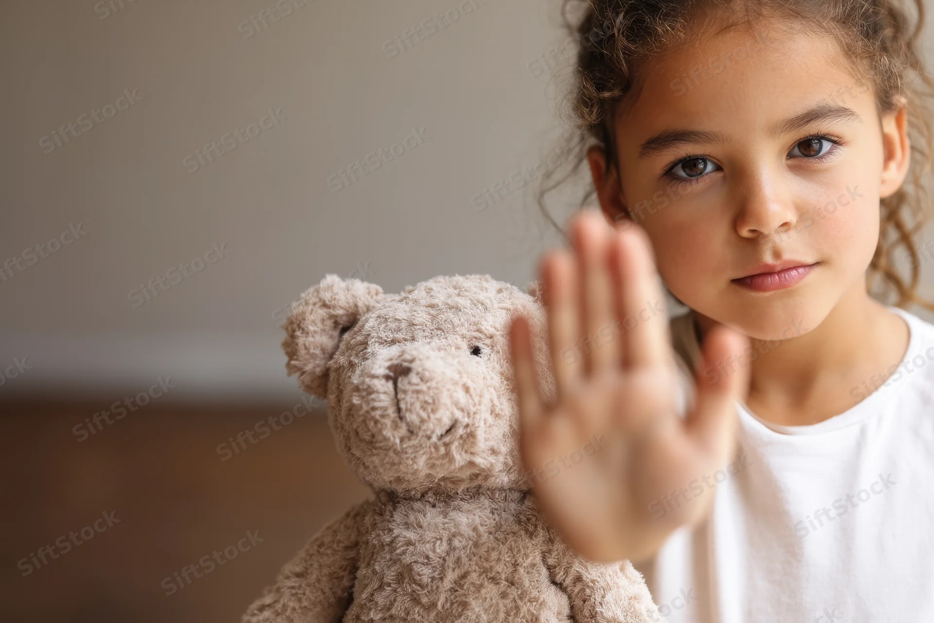 Young girl holding teddy bear and raising hand in stop gesture indoors