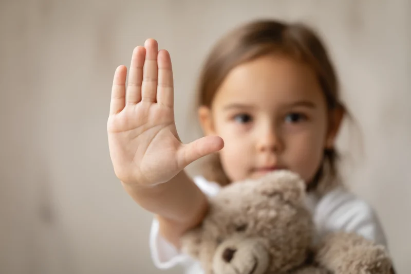 Young girl holding teddy bear and raising hand in stop gesture indoors