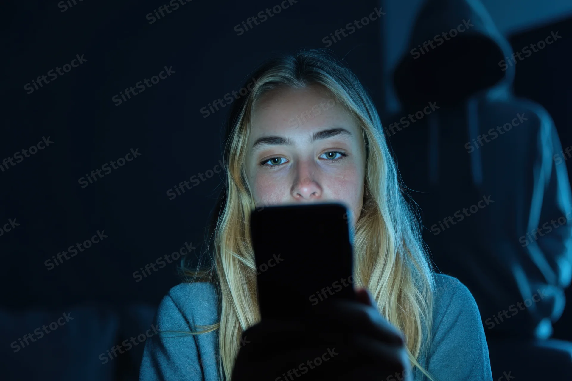 Young girl illuminated by smartphone screen in dark room holding device with hooded figure in background