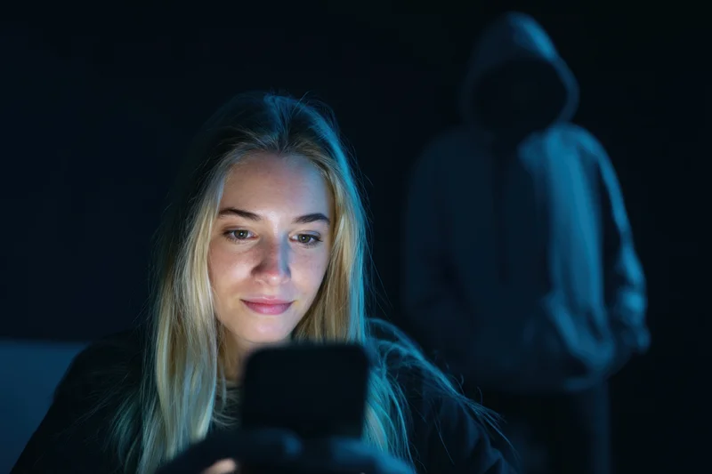 Young woman illuminated by smartphone screen in dark room with shadowy hooded figure behind her