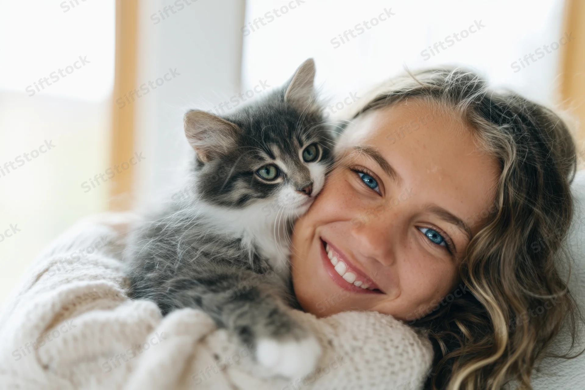 Young woman smiling while embracing a fluffy gray kitten wrapped in a knit blanket indoors