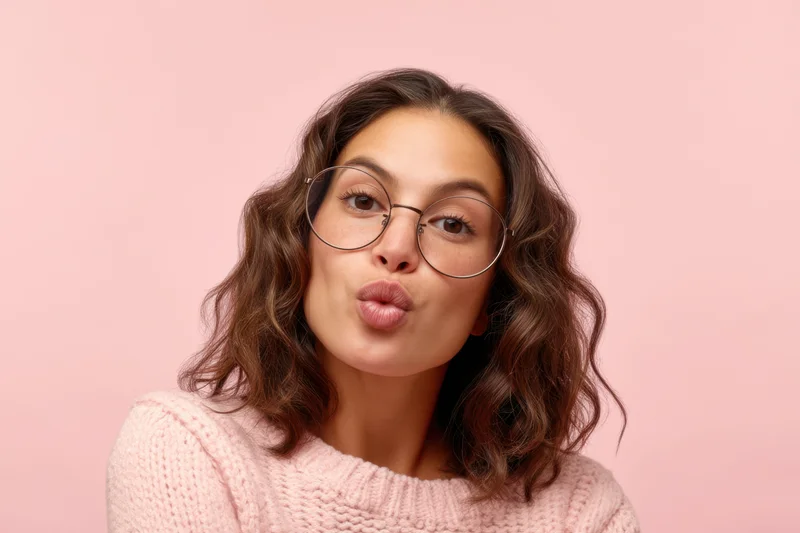 Young woman making playful pouting expression with round glasses and pink knit sweater against pink background