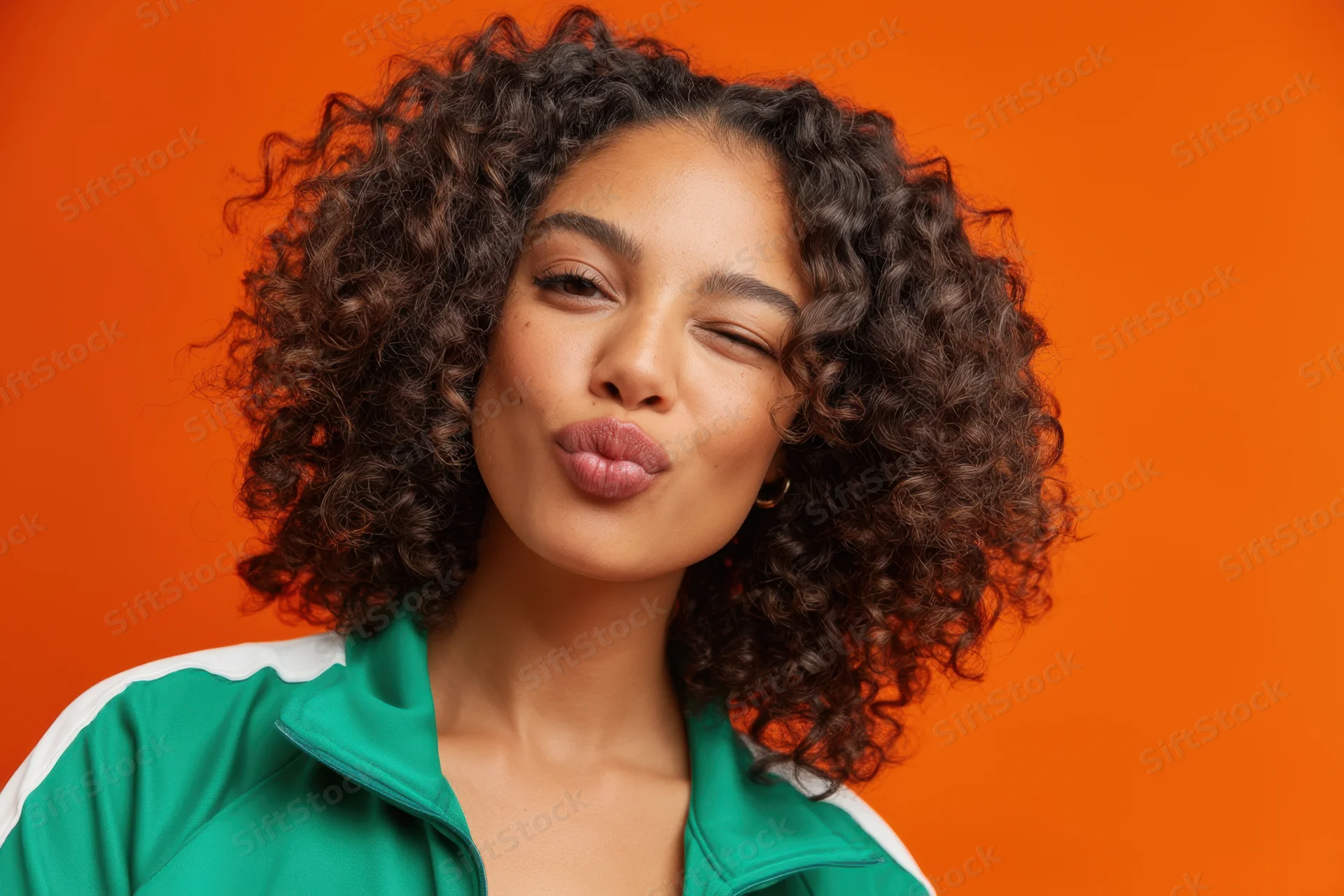 Young black woman with curly hair making playful wink against orange background wearing green jacket with white stripe