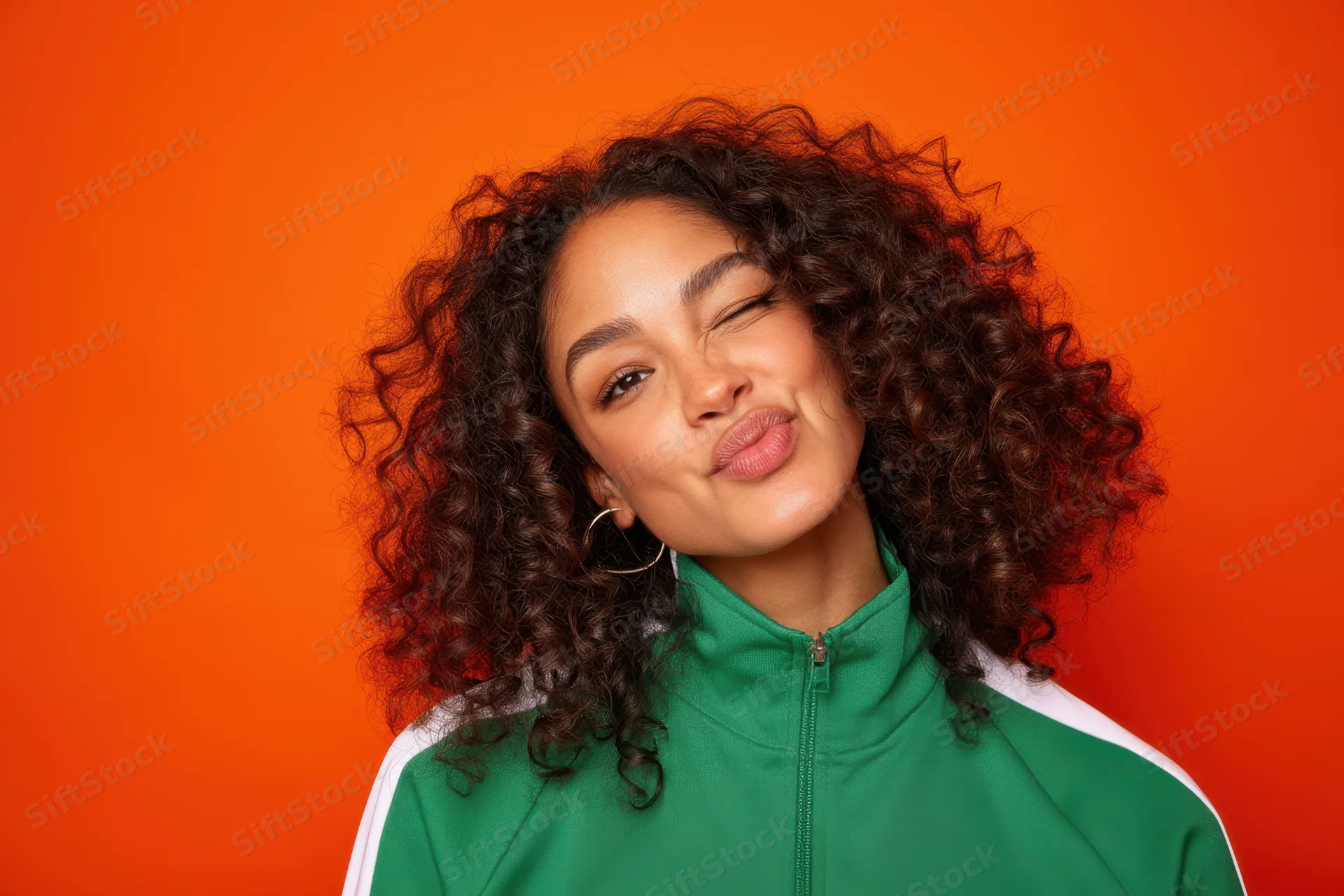 Young woman with curly hair winking playfully against orange background wearing green tracksuit