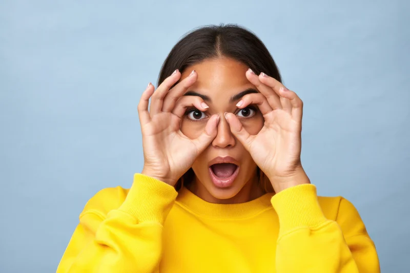 Young woman making binoculars gesture with hands over eyes in yellow sweatshirt against blue background