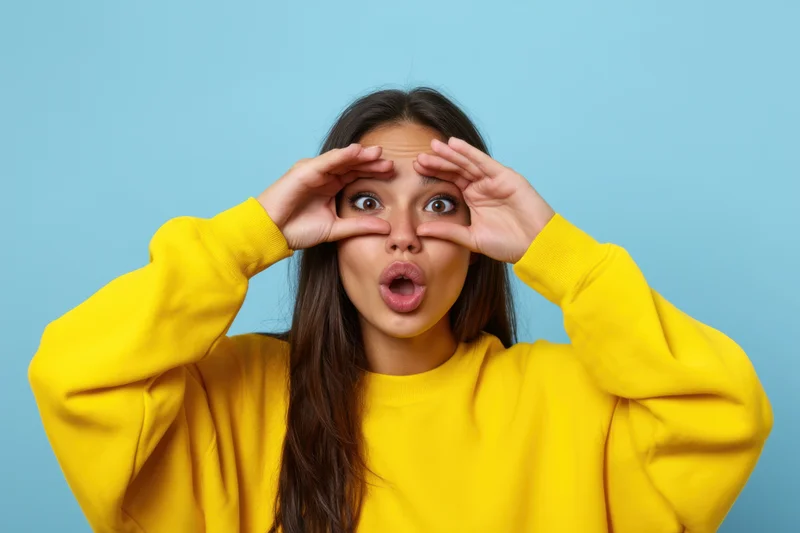 Young woman making binoculars gesture with hands over eyes in yellow sweatshirt against blue background