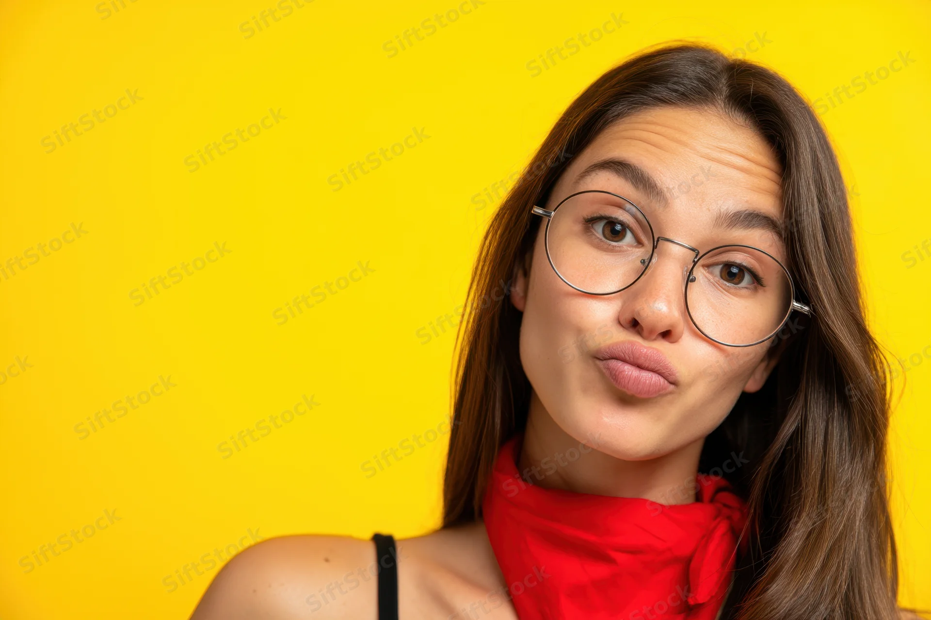 Young woman with long brown hair and round glasses making a playful pout against yellow background