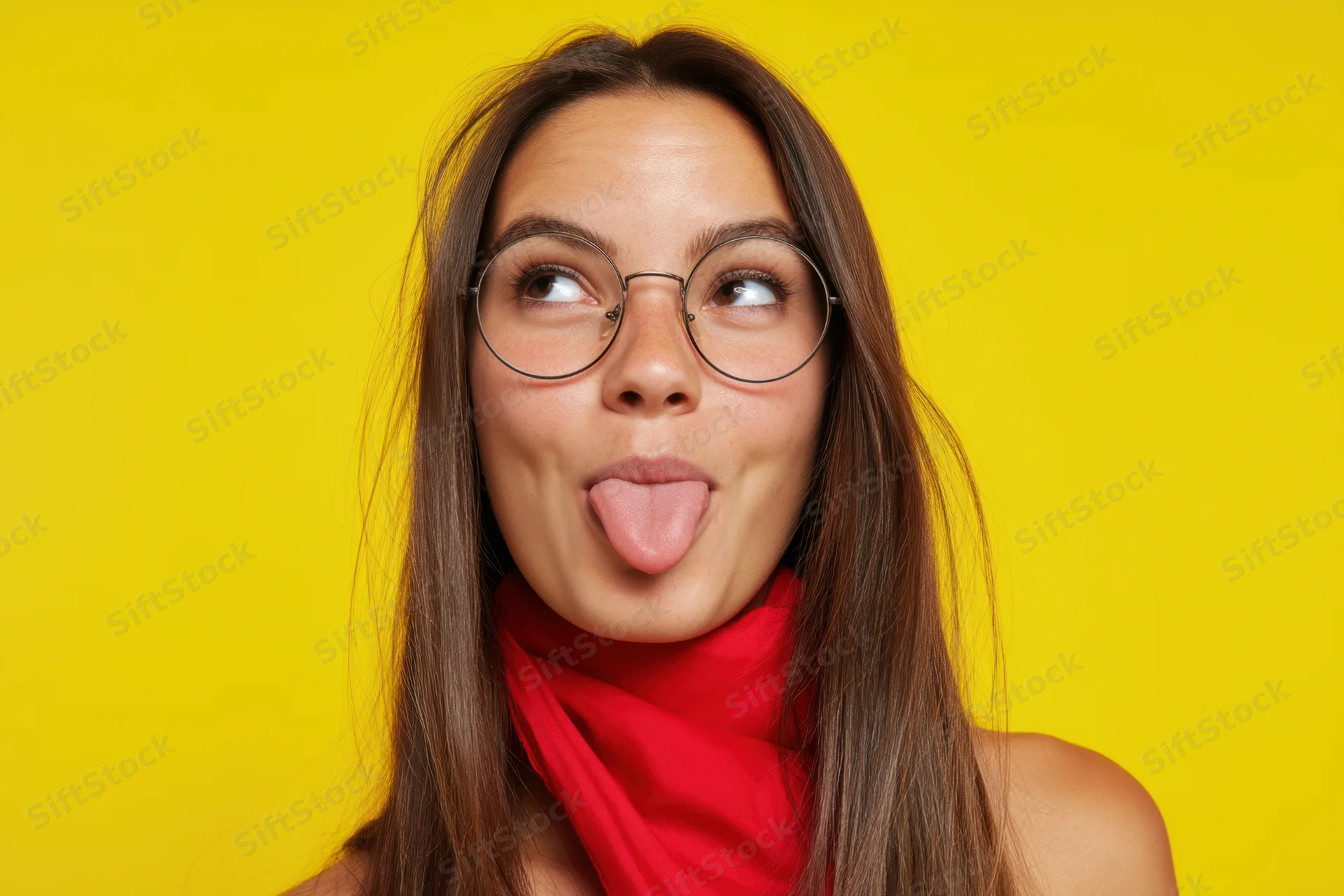 Young woman with long brown hair sticking out her tongue while wearing round glasses and a red scarf against a yellow background